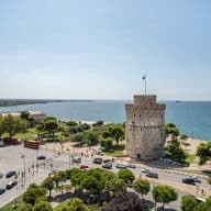 Ένα παραθαλάσσιο αστικό τοπίο της Θεσσαλονίκης παρουσιάζει τον εμβληματικό Λευκό Πύργο δίπλα στη θάλασσα, που περιβάλλεται από δέντρα και πολυσύχναστους δρόμους με αυτοκίνητα και λεωφορεία. Ο ουρανός είναι καθαρός και ηλιόλουστος, ενώ η απεριόριστη θάλασσα απλώνεται στον ορίζοντα.