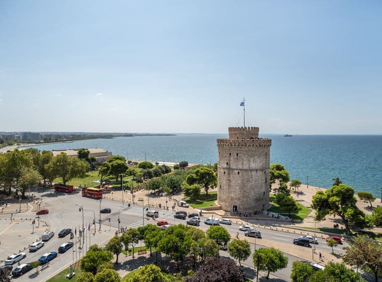 Ένα παραθαλάσσιο αστικό τοπίο της Θεσσαλονίκης παρουσιάζει τον εμβληματικό Λευκό Πύργο δίπλα στη θάλασσα, που περιβάλλεται από δέντρα και πολυσύχναστους δρόμους με αυτοκίνητα και λεωφορεία. Ο ουρανός είναι καθαρός και ηλιόλουστος, ενώ η απεριόριστη θάλασσα απλώνεται στον ορίζοντα.