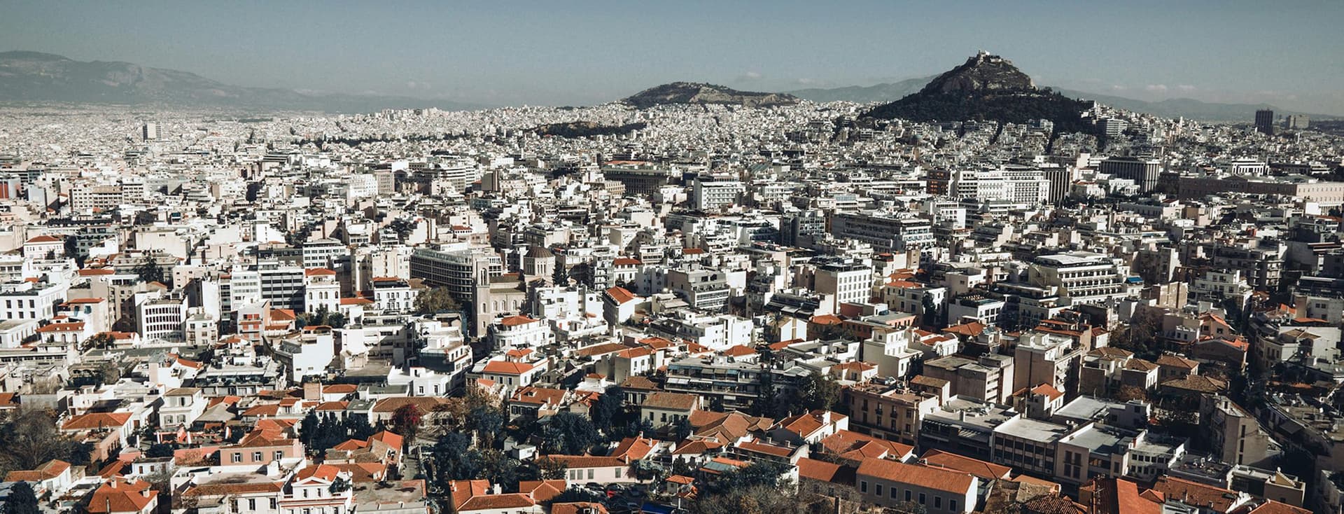 Αεροφωτογραφία της Αθήνας, που παρουσιάζει ένα τεράστιο αστικό τοπίο με πυκνά γεμάτα κτίρια, στέγες με κόκκινα κεραμίδια και το όρος Λυκαβηττό στο βάθος κάτω από έναν καταγάλανο ουρανό.
