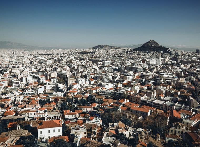 Αεροφωτογραφία της Αθήνας, που παρουσιάζει ένα τεράστιο αστικό τοπίο με πυκνά γεμάτα κτίρια, στέγες με κόκκινα κεραμίδια και το όρος Λυκαβηττό στο βάθος κάτω από έναν καταγάλανο ουρανό.