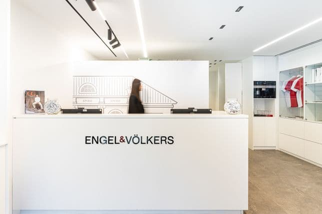 Modern office reception with a white desk displaying "Engel & Völkers" logo. A person walks by, and shelves with books and awards are in the background.