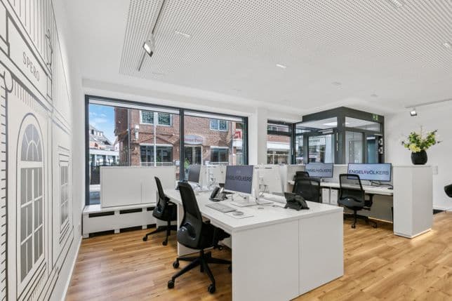 Modern office space with wooden floors, white desks, black chairs, and large windows. A wall mural and a vase with flowers are visible.