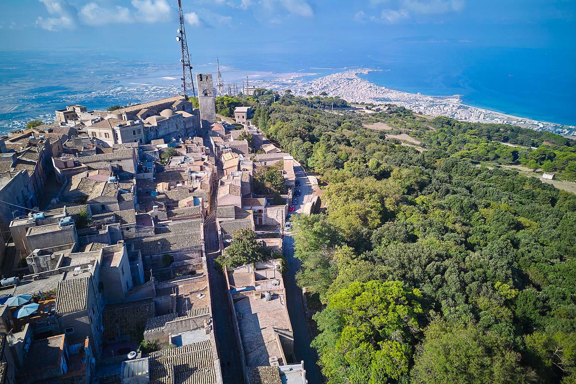 Luftaufnahme einer historischen Stadt am Hang mit Steinhäusern, einem Turm und üppigem Grün mit Blick auf eine Küstenstadt und das blaue Meer im Hintergrund.