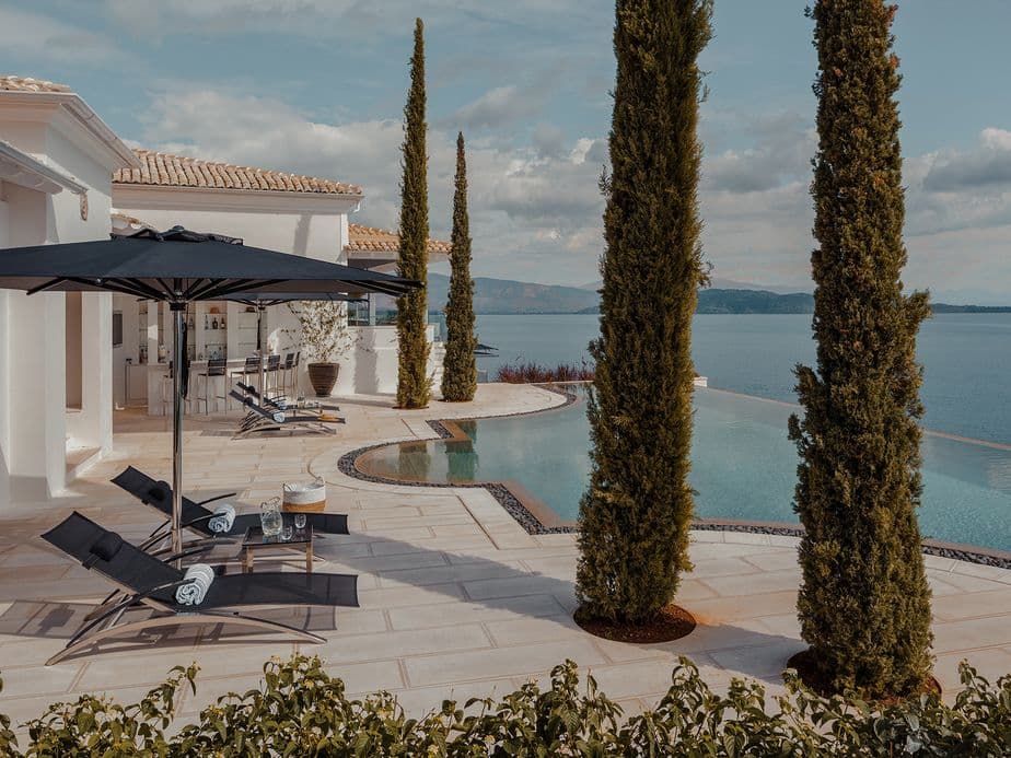A luxurious seaside villa with an infinity pool, lounge chairs, umbrellas, and tall cypress trees overlooking a calm blue sea under a partly cloudy sky.