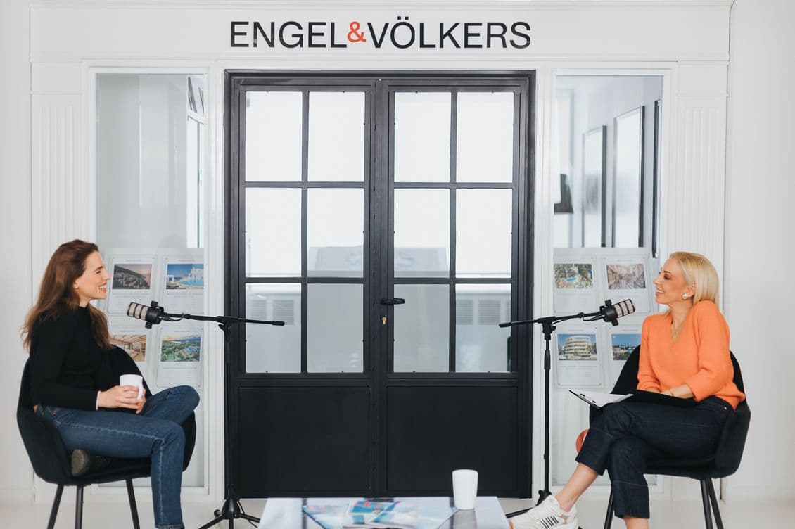 Two women sit with microphones in front of a door labeled "ENGEL & VÖLKERS," engaged in conversation, one holding a cup, the other a notebook.