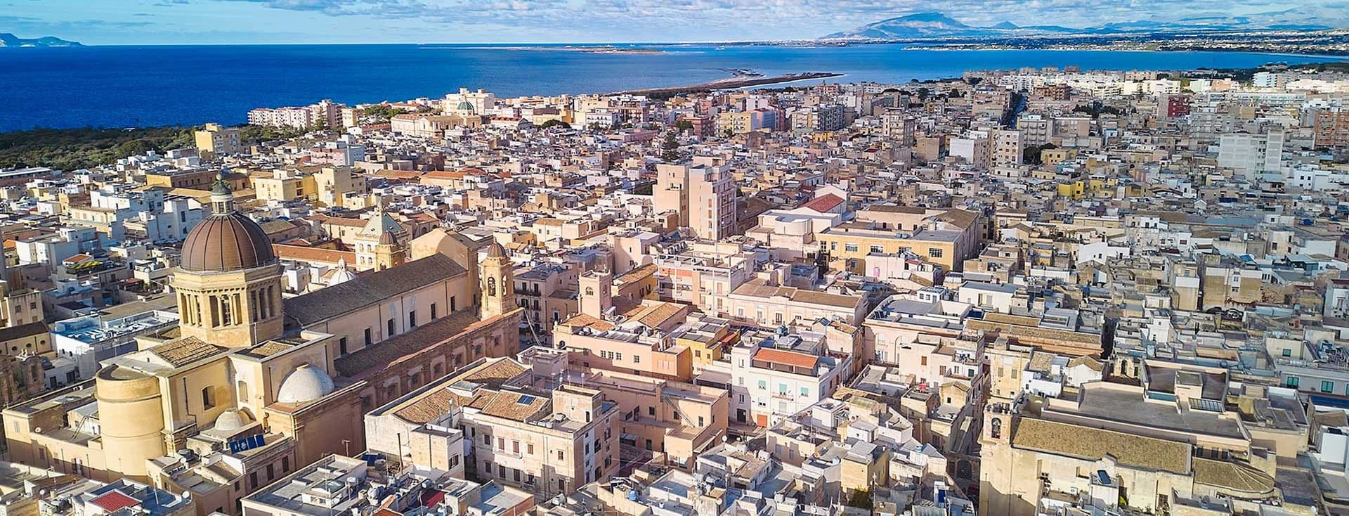 Aerial view of a coastal city with a prominent dome-shaped church, dense buildings, and the sea in the background under a partly cloudy sky.
