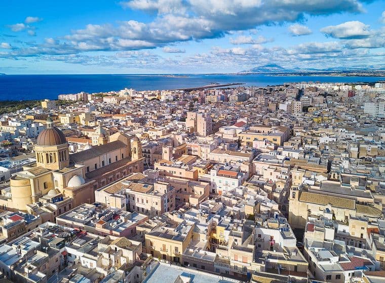 Luftaufnahme einer Küstenstadt mit einer markanten kuppelförmigen Kirche, dichter Bebauung und dem Meer im Hintergrund unter einem teilweise bewölkten Himmel.