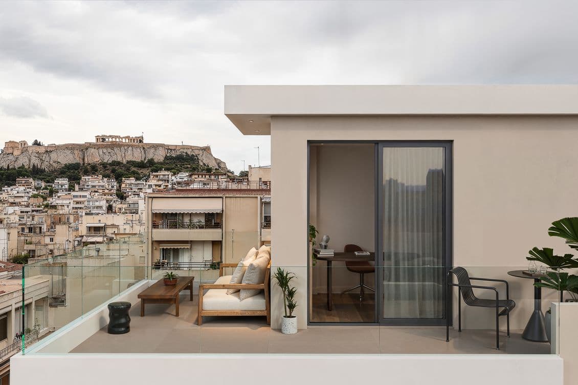 Modern rooftop terrace with beige sofa, coffee table, and plant. Glass railing overlooks cityscape and Acropolis under cloudy sky.