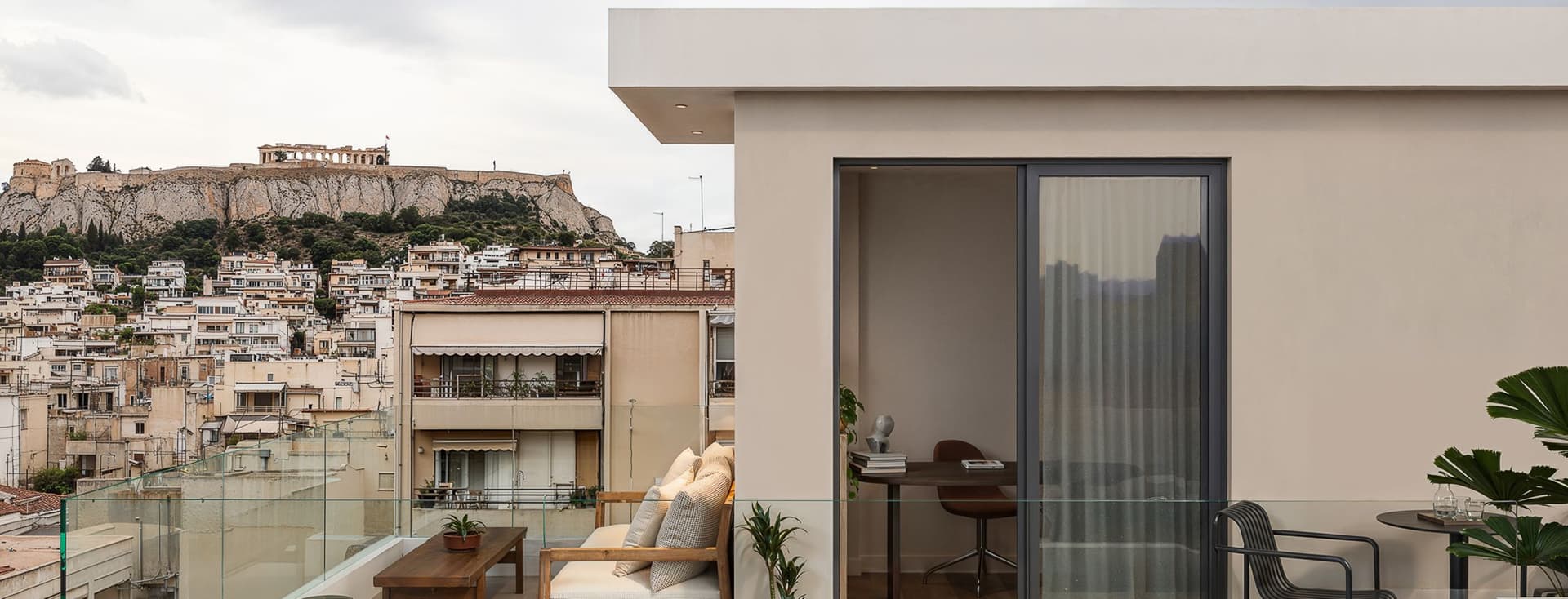Modern rooftop terrace with beige sofa, coffee table, and plant. Glass railing overlooks cityscape and Acropolis under cloudy sky.