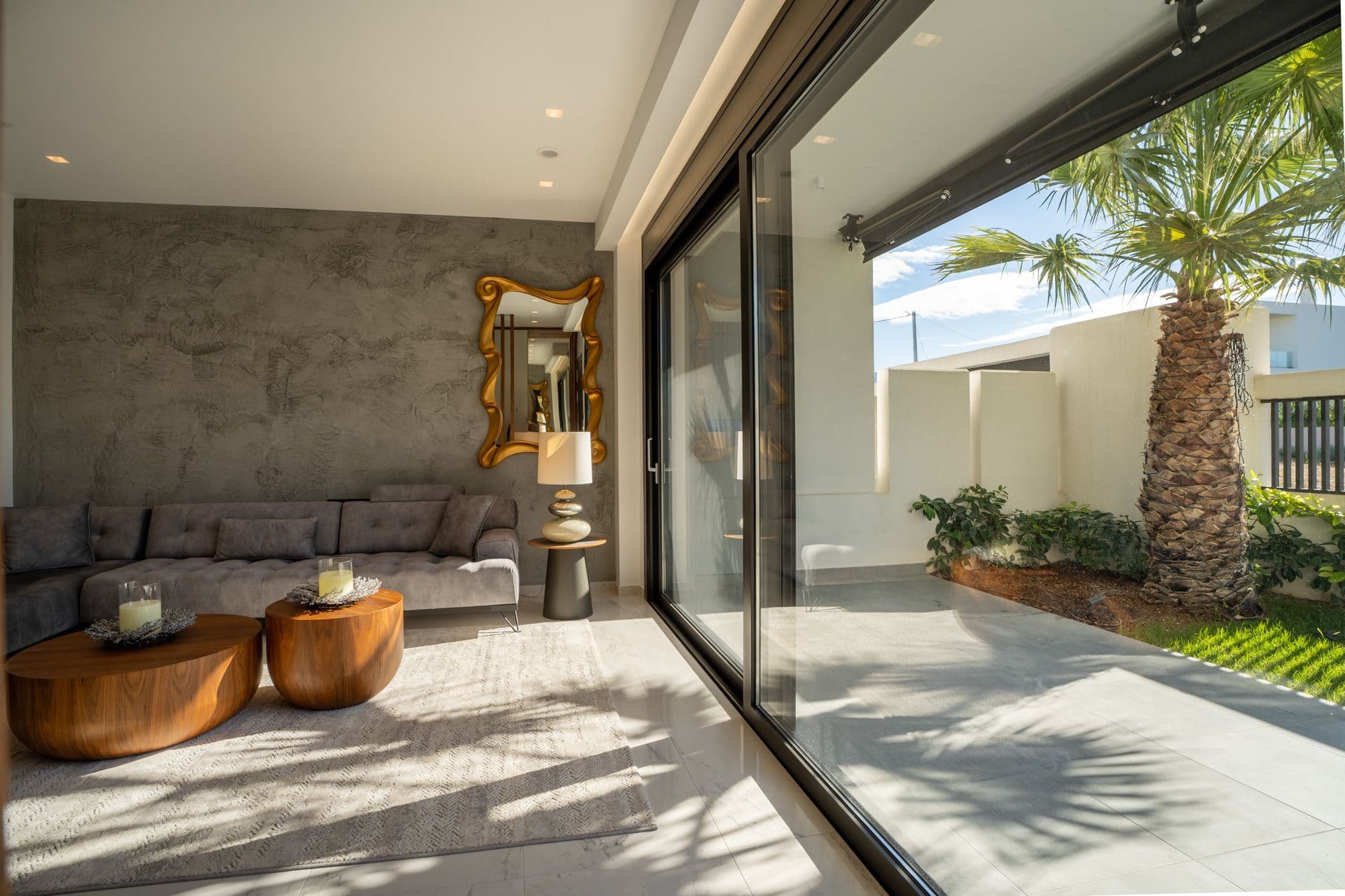 Modern living room with gray sofa, wooden coffee tables, and large glass doors opening to a sunny patio with a palm tree and greenery.