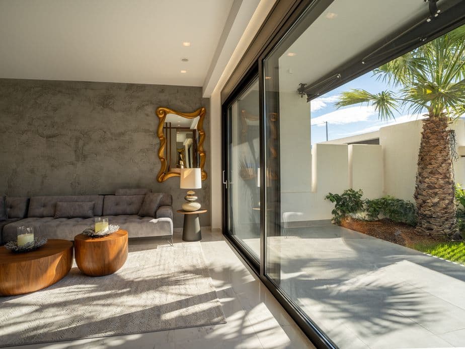 Modern living room with gray sofa, wooden coffee tables, and large glass doors opening to a sunny patio with a palm tree and greenery.