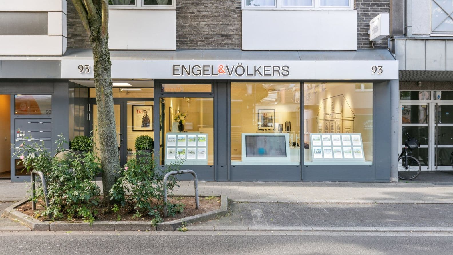 Storefront of Engel & Völkers with large windows displaying real estate listings, a tree on the sidewalk, and a bicycle parked nearby.