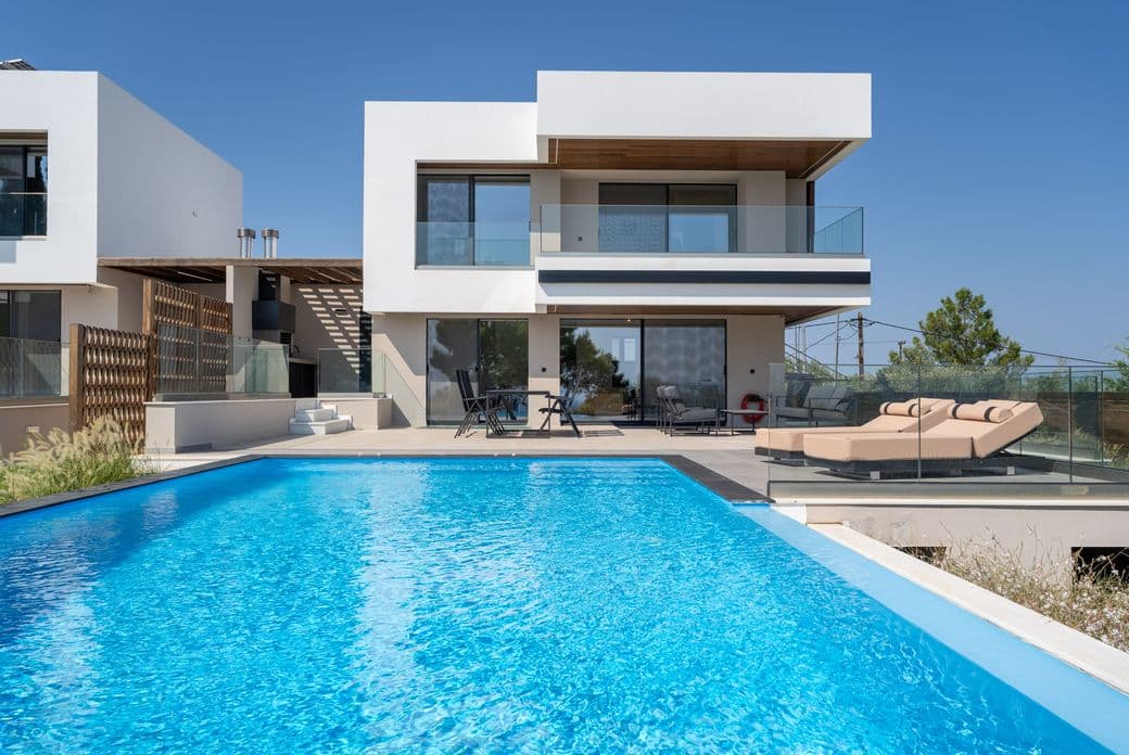 Modern two-story house with a glass balcony, pool, and sun loungers on a clear day. Minimalist design with white and wood accents.