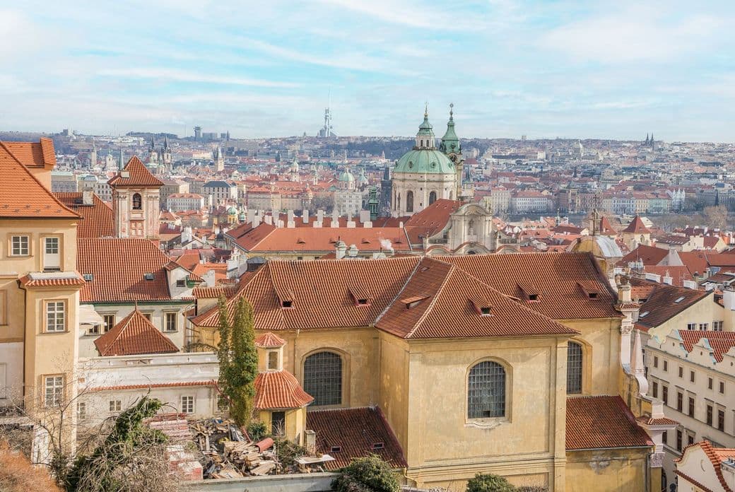 Elevated view of Prague’s Lesser Town with red rooftops, Baroque churches, and the prominent dome of St. Nicholas Church. In the background, the city skyline with the Žižkov TV Tower and Prague Castle.
