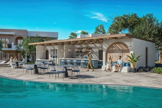 Outdoor poolside area with people relaxing, shaded seating, and a bar under a pergola. Trees and clear blue sky in the background.