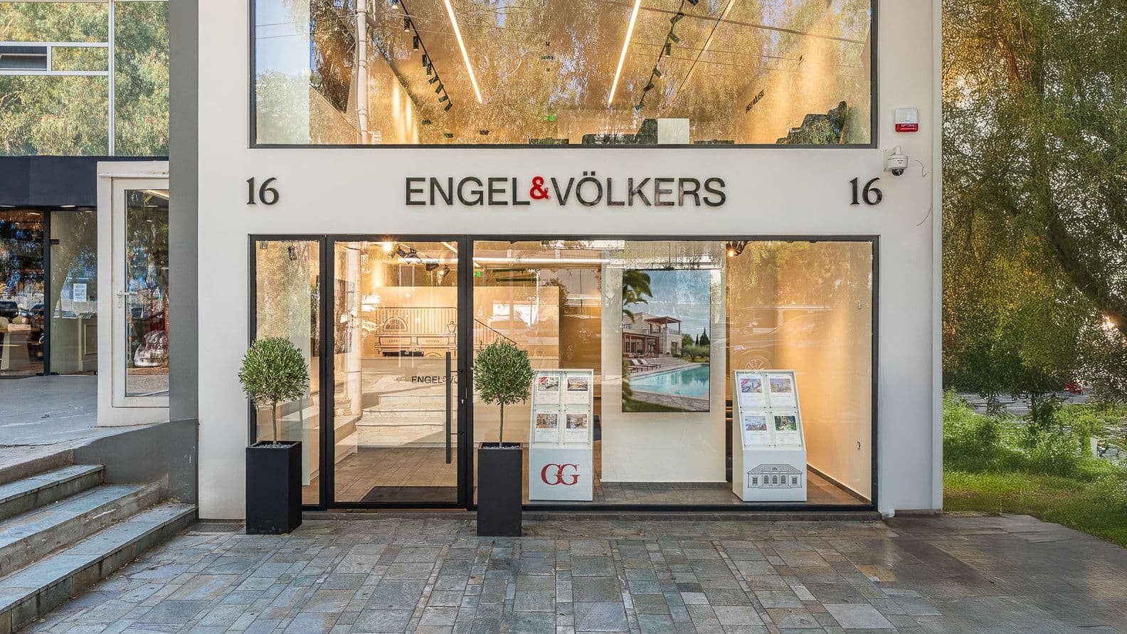 Modern real estate office with large glass windows displaying property listings, two decorative potted plants flanking the entrance.