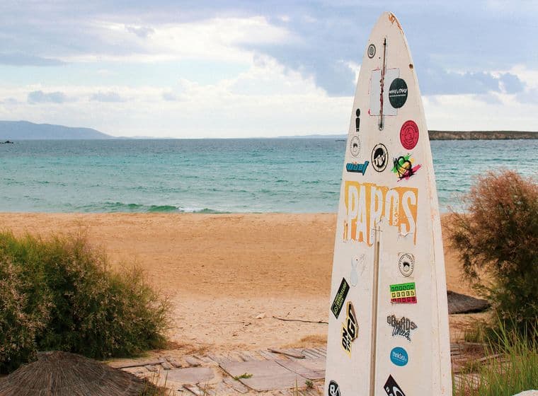 Μια αμμώδης παραλία στην Πάρο οδηγεί σε ήρεμη γαλάζια θάλασσα κάτω από έναν συννεφιασμένο ουρανό. Στο πρώτο πλάνο, μια σανίδα του σερφ καλυμμένη με πολύχρωμα αυτοκόλλητα στέκεται όρθια κοντά σε μερικούς θάμνους.