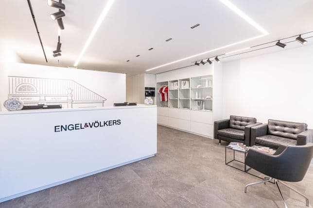 Modern office lobby with a reception desk labeled "Engel & Völkers," two black armchairs, a magazine table, and a decorative shelf.