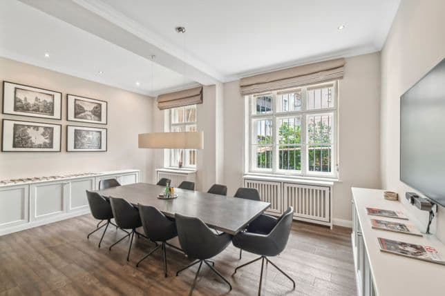 Ein modernes Esszimmer mit einem langen Tisch, schwarzen Stühlen, großen Fenstern, gerahmten Bildern an der Wand und einem Fernseher auf einem weißen Schrank.