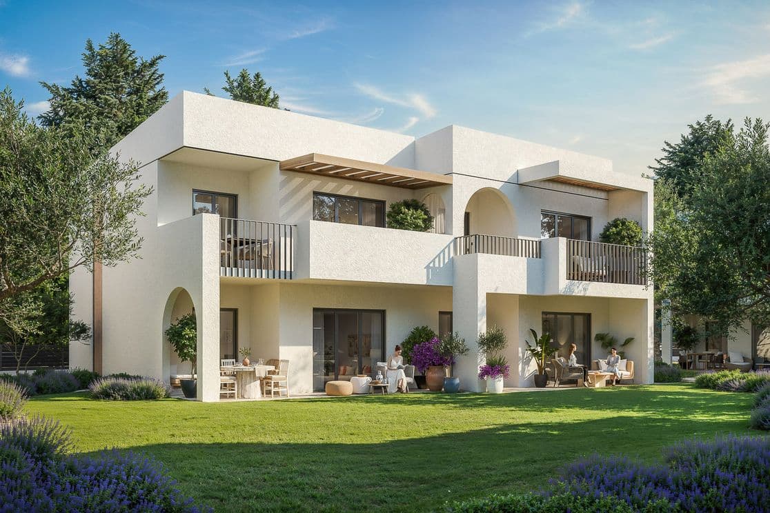 Modern white two-story house with balconies, large windows, patio seating, surrounded by lush greenery and trees under a clear blue sky.