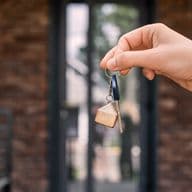 Hand holding a key with a house-shaped keychain in front of a brick building entrance.