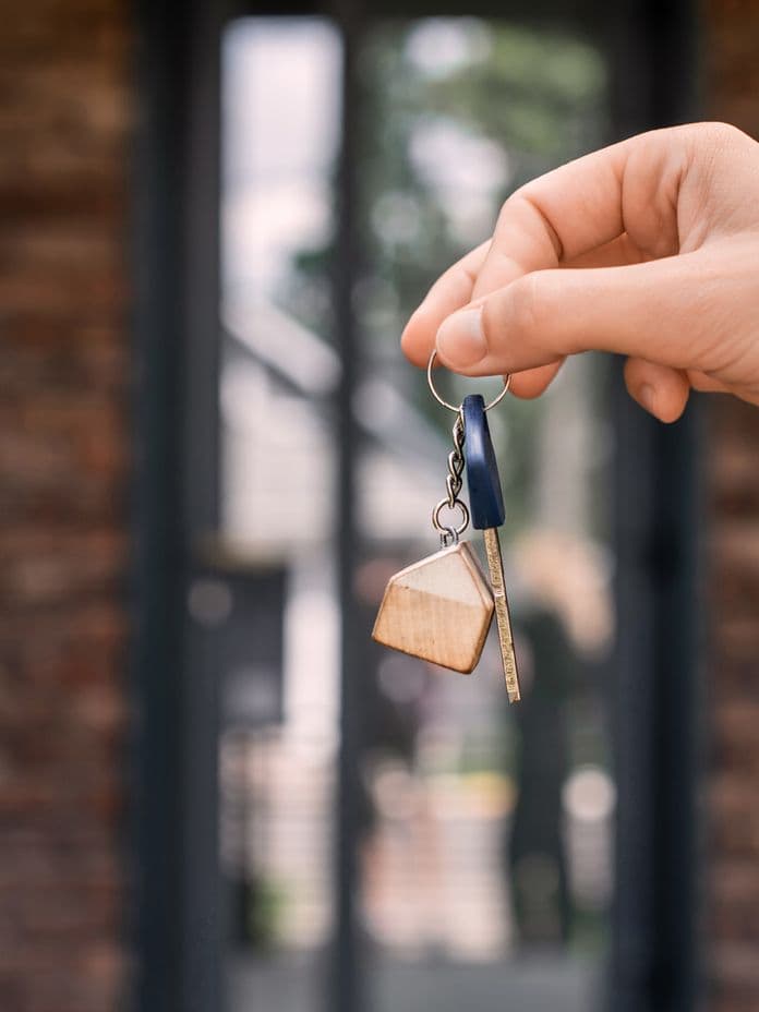 Hand holding a key with a house-shaped keychain in front of a brick building entrance.