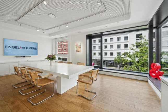 Moderner Konferenzraum mit einem langen weißen Tisch, Holzstühlen, einem an der Wand befestigten Bildschirm, großen Fenstern und Holzboden.