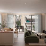 Modern living room with a green sofa, coffee table, and wall-mounted TV, leading to a balcony with outdoor seating. Natural light fills the space.