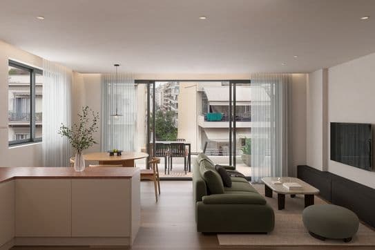 Modern living room with a green sofa, coffee table, and wall-mounted TV, leading to a balcony with outdoor seating. Natural light fills the space.