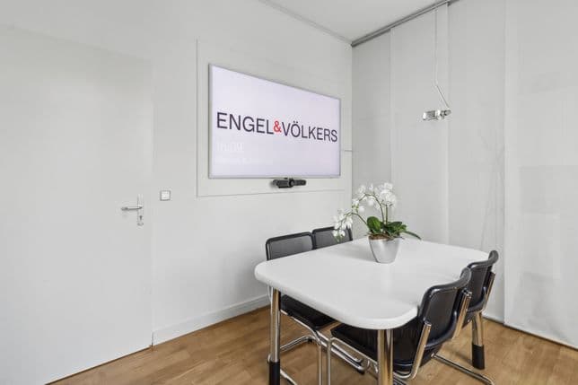 Minimalistischer Besprechungsraum mit weißem Tisch, schwarzen Stühlen, einer getopften Orchidee und einem an der Wand befestigten Bildschirm mit der Aufschrift „ENGEL & VÖLKERS“.