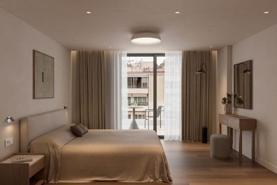 Minimalist bedroom with a large bed, wooden floor, and a window with sheer curtains. A small desk, chair, and wall art complete the decor.
