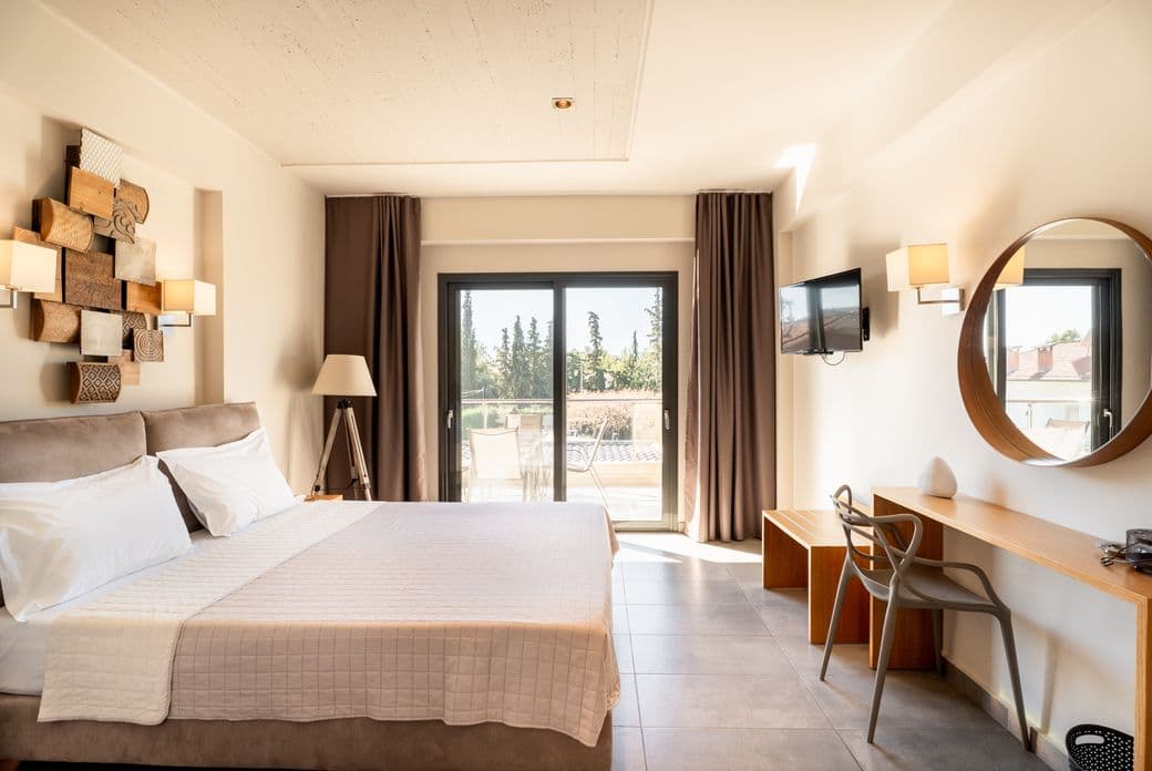 Modern hotel room with a double bed, wall art, desk, chair, round mirror, TV, and large window leading to a balcony with outdoor views.