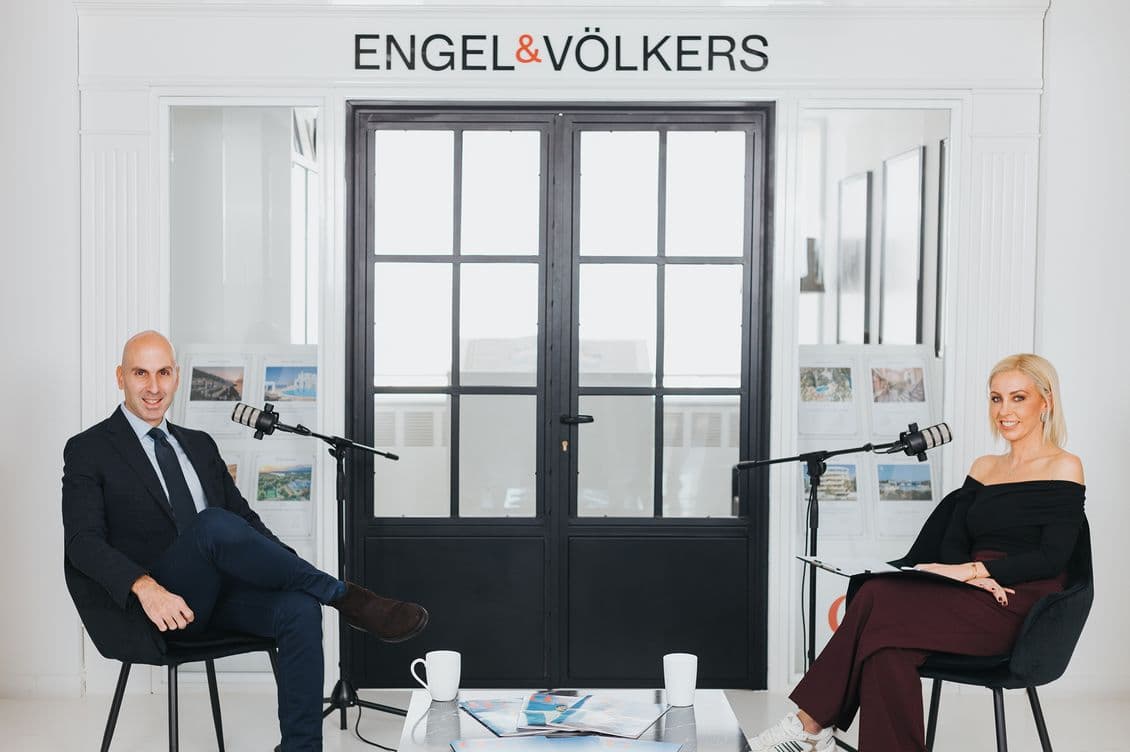 Two people seated with microphones in a modern office setting, under the sign "Engel & Völkers." A table with papers and mugs is in front.