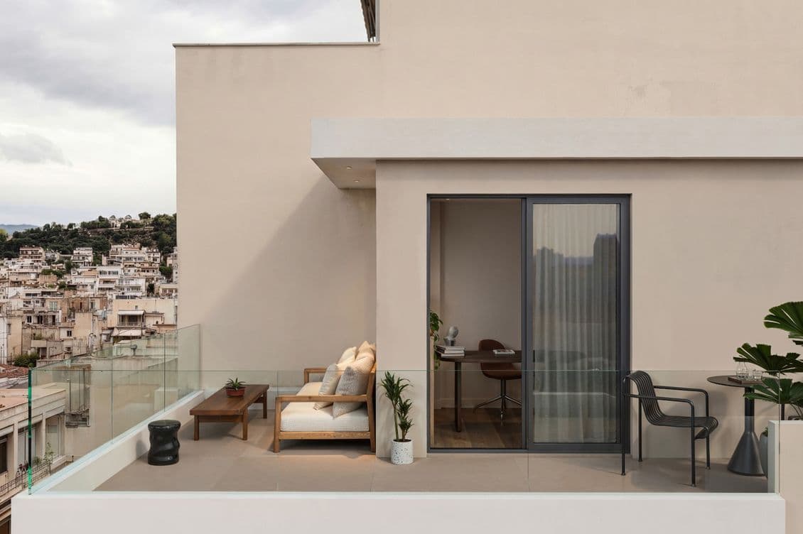 Modern rooftop patio with glass railings, beige walls, a cozy sofa, potted plants, and a sliding door leading to an interior office space.