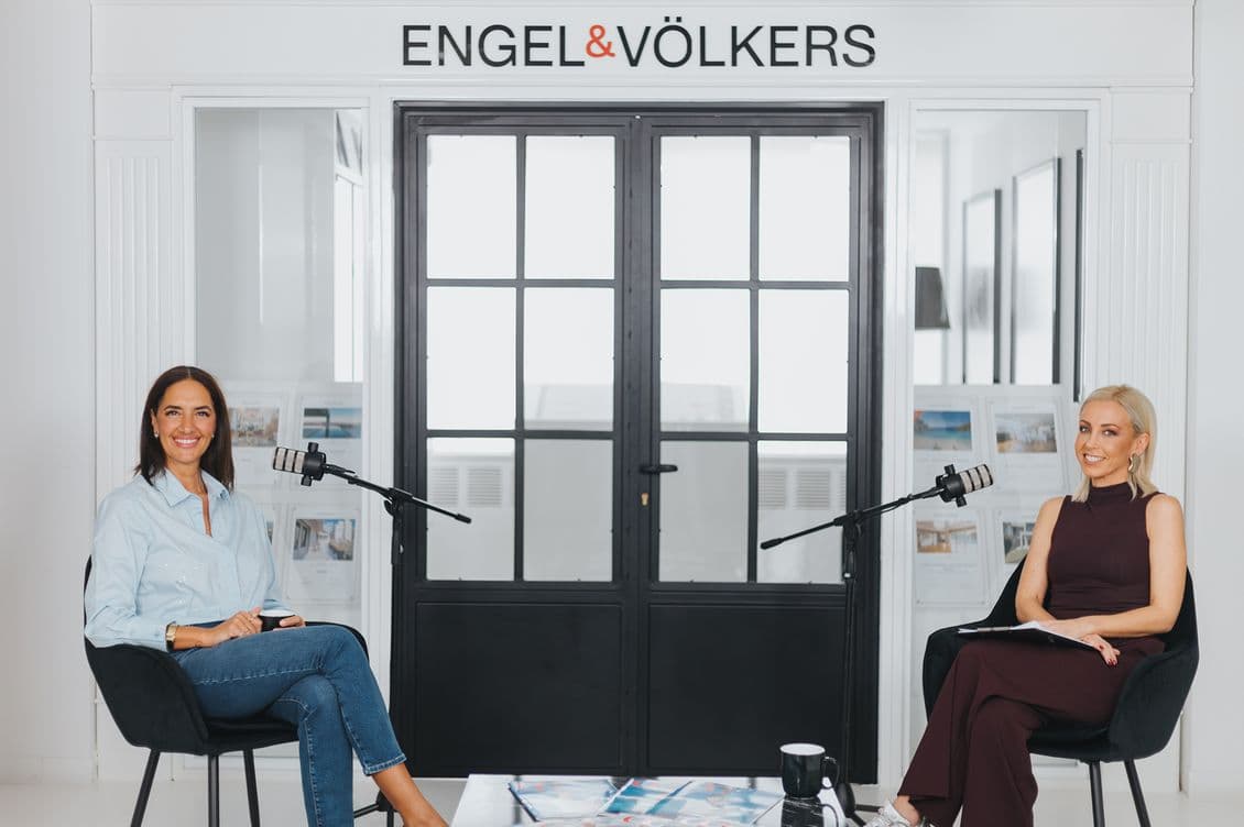 Two women sitting with microphones in an office setting, engaged in a discussion. Background features "Engel & Völkers" signage.