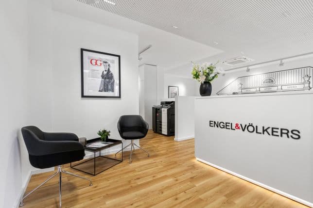 Modern office lobby with two black chairs, a framed photo on the wall, a printer, and a reception desk with "Engel & Völkers" logo.
