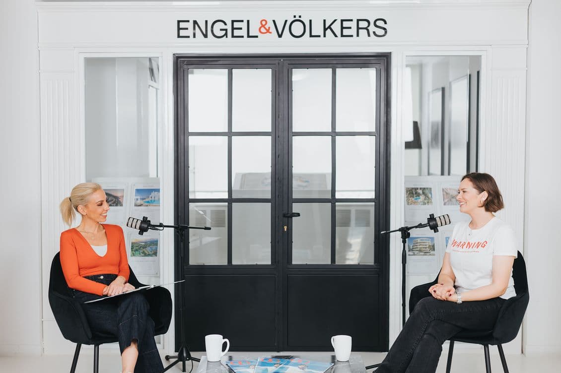 Two women in casual attire sit facing each other, discussing in a bright room with microphones, Engel & Völkers sign above.