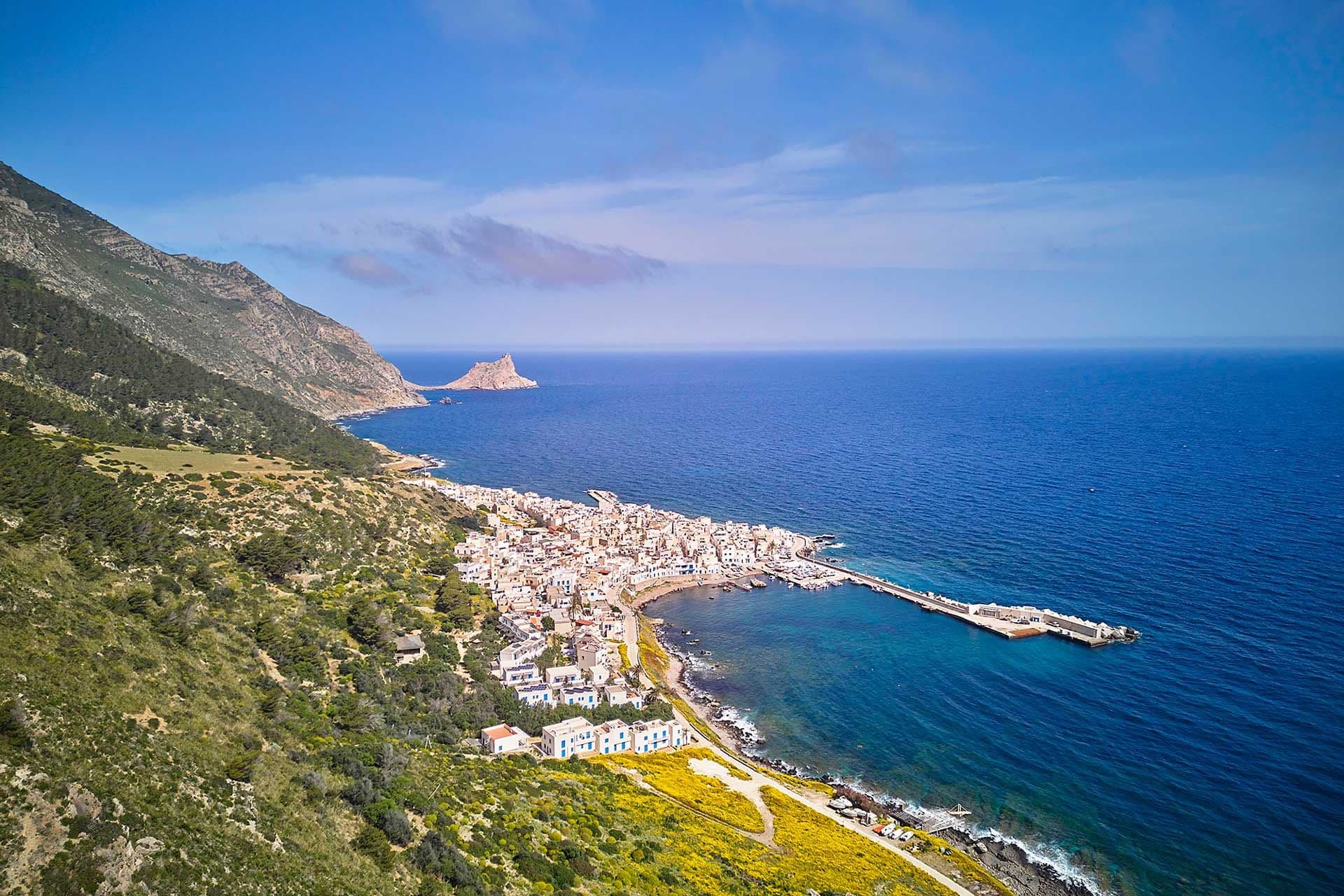 Veduta aerea di una città costiera con un porto, circondata da una vegetazione lussureggiante e da un paesaggio montuoso, sotto un cielo azzurro e limpido.