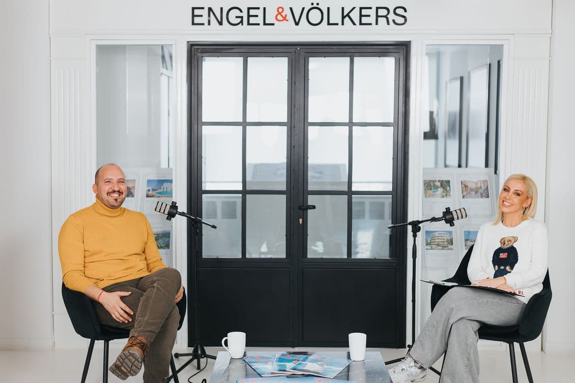 Two people seated in a modern office setting with microphones, smiling at the camera. A sign above reads "Engel & Völkers."
