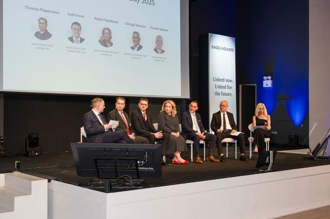 Eight people seated on stage in a panel discussion setting, with a man speaking into a microphone. A screen displays names and roles above them.