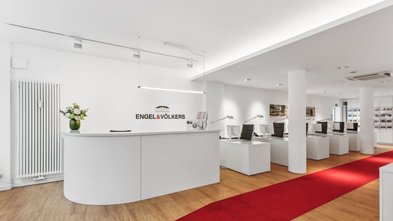 Moderner Büroempfang mit weißen Schreibtischen, rotem Teppich und Computern. Eine Vase mit Blumen steht auf dem Tresen unter heller Deckenbeleuchtung.