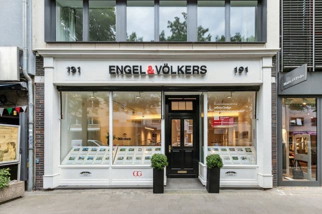 Schaufenster von Engel & Völkers mit großen Schaufenstern, die Immobilienangebote anzeigen, einer schwarzen Tür und zwei Topfpflanzen.