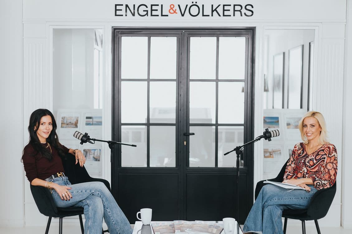 Δύο γυναίκες, η Λιλή Πυράκη και η Ξένια Κωνσταντίνου, κάθονται η μία απέναντι από την άλλη σε ένα σύγχρονο γραφείο με μικρόφωνα, φλιτζάνια καφέ και περιοδικά στο τραπέζι. Πίσω τους, μεγάλα παράθυρα πλαισιώνουν τις λέξεις ENGEL & VÖLKERS πάνω από τις πόρτες.
