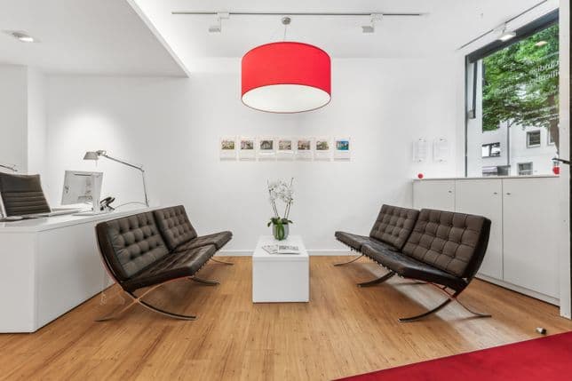 Modernes Wartezimmer mit zwei dunklen Ledersofas, einem weißen Tisch mit einer Pflanze und einer roten Deckenlampe. Großes Fenster und gerahmte Bilder an der Wand.