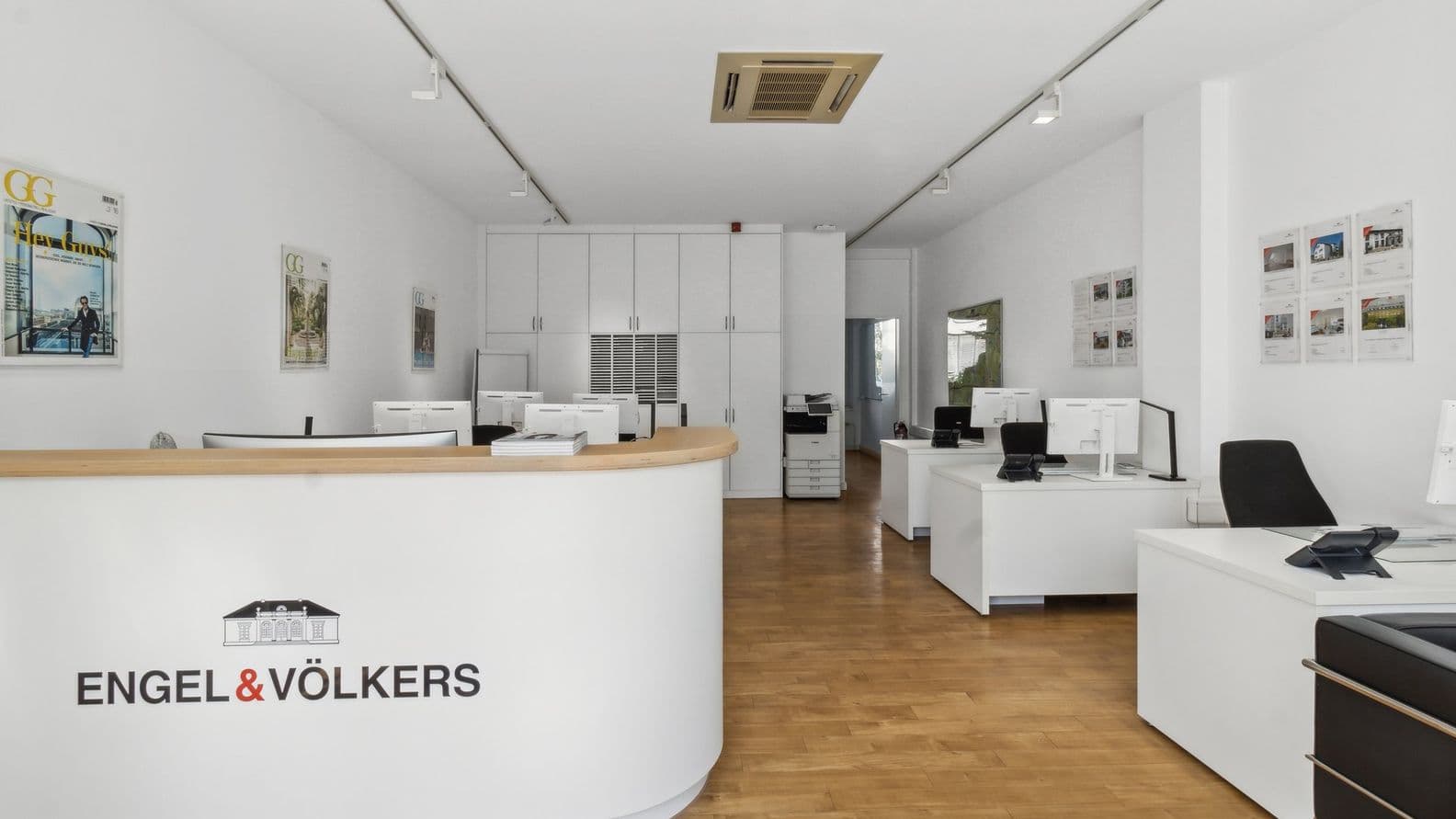 Minimalistischer Besprechungsraum mit weißem Tisch, schwarzen Stühlen, einer getopften Orchidee und einem an der Wand befestigten Bildschirm mit der Aufschrift „ENGEL & VÖLKERS“. Modernes Immobilienbüro mit weißen Schreibtischen, schwarzen Stühlen und einem gebogenen Empfangstresen. An den Wänden hängen gerahmte Bilder und Branding.