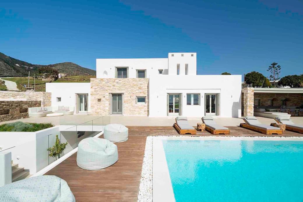 Modern white villa with stone accents, multiple windows, and a flat roof, featuring a wooden pool deck, light-colored bean bags, lounge chairs, and a turquoise swimming pool under a clear blue sky.