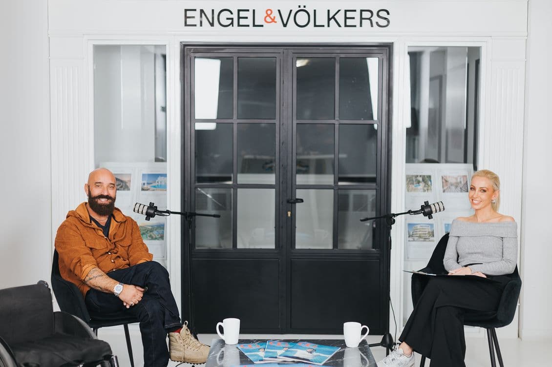Two people seated with microphones in an Engel & Völkers studio, engaged in a discussion. A table with cups and magazines is in the foreground.