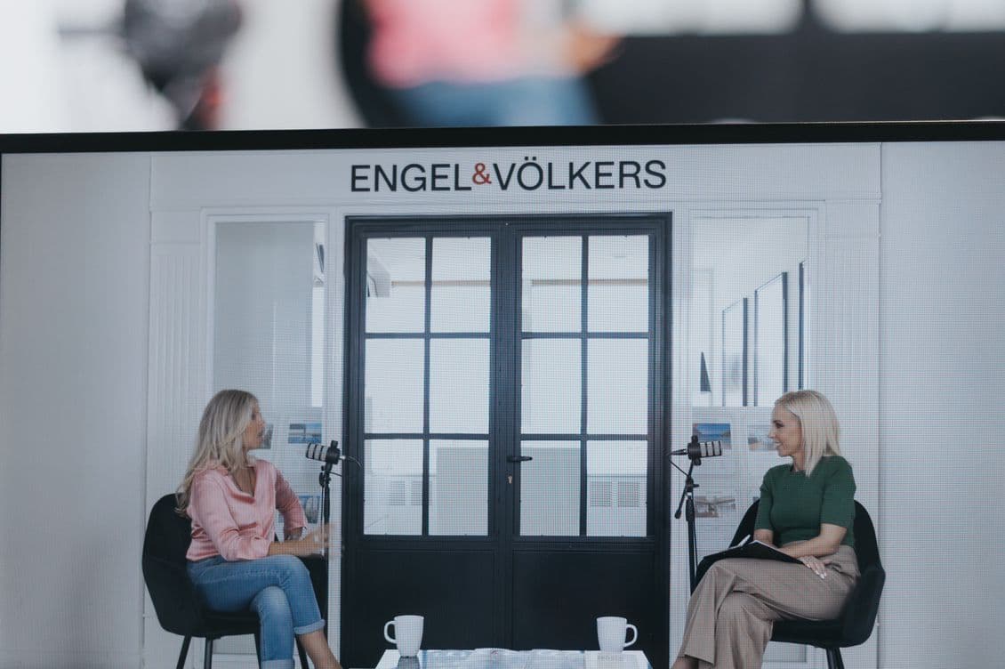 Two women sit in chairs facing each other in a modern office setting with microphones, under a sign reading "ENGEL & VÖLKERS."