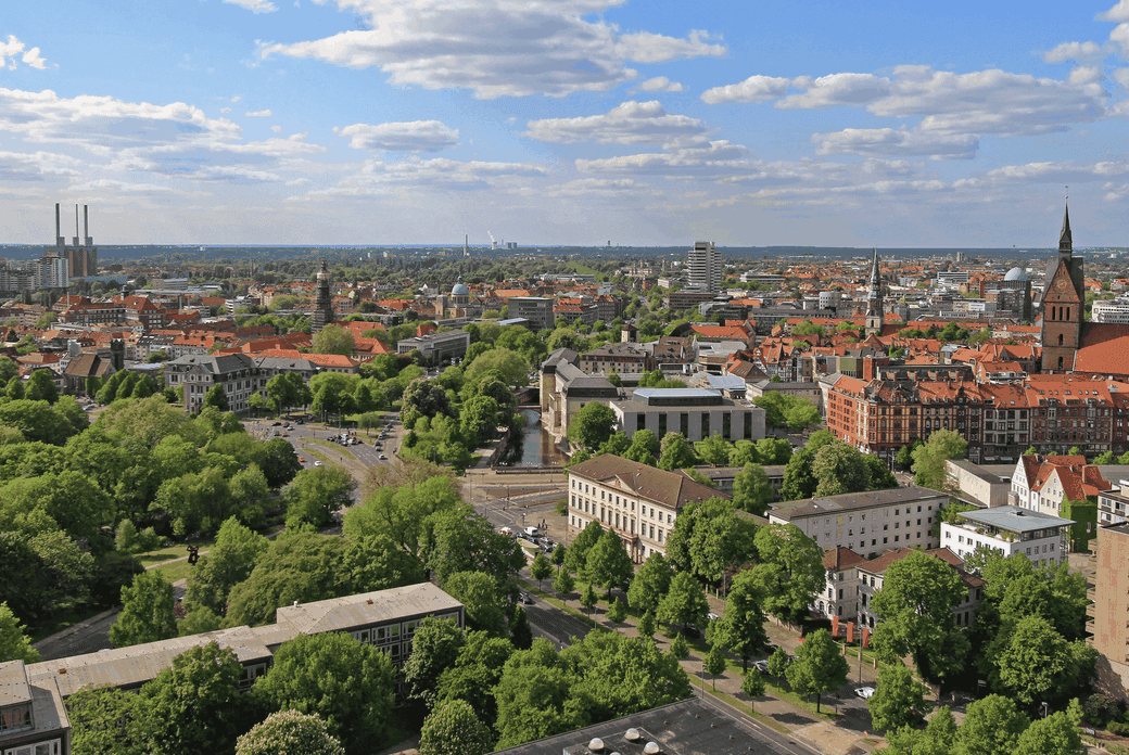Aerial view of a city with historic buildings, green trees, and a clear blue sky with scattered clouds.