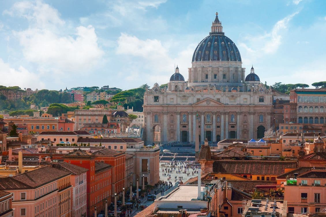 Aerial view of St. Peter's Basilica in Vatican City, surrounded by historic buildings and a bustling square under a blue sky.